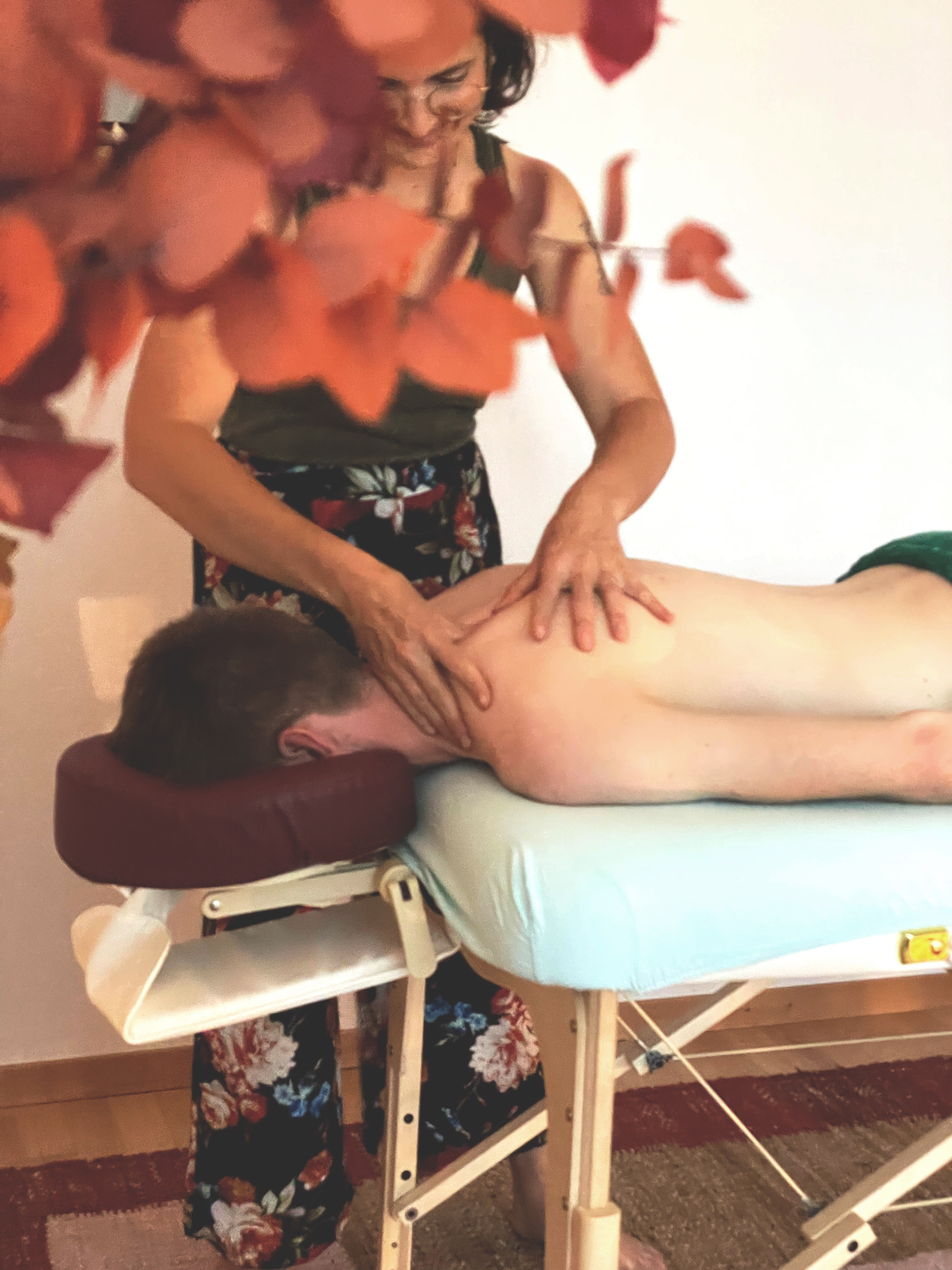 Photo d'une démonstration d'un massage. La thérapeute Camille Létang se tient debout avec le sourire, les mains en action sur la peau de l'omoplate d'une personne allongée sur le ventre.